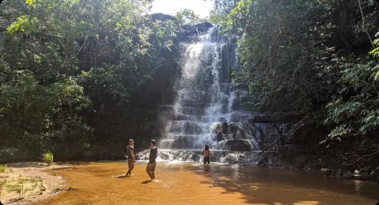 uma das cachoeiras da trilha. Existe 4 pessoas na foto. A cachoeira tem mais de 5 metros de altura e possui pedras que se assemelham a grandes degraus. A Agua está com um alaranjado fraco.